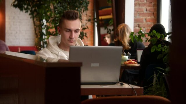 Tired young man is working on laptop in cafe. Stock footage. Handsome young man is writing diploma on laptop in cafe. Young freelancer or student is working on laptop in cafe