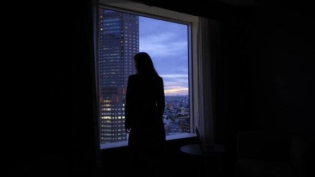 Woman come to window and look out to evening city, dark room, silhouetted figure against window, view from back and side. Lady wear informal suit. Blurred Tokyo cityscape and tall tower on back