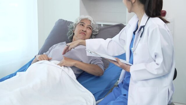 Elderly Asian woman lying in hospital bed looking unwell, with young Asian doctor take a history of illness Health check and encouragement.