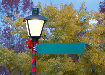Close up on an Old Fashioned Street Light burning at sunset, covered in red ribbon and bow for Christmas. Street sign blank for your text.