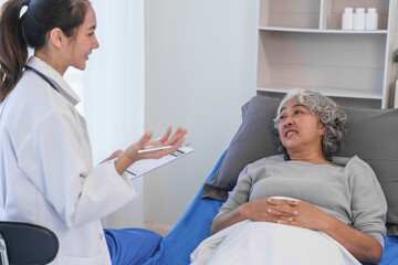 Elderly Asian woman lying in hospital bed looking unwell, with young Asian doctor take a history of...