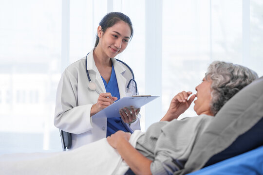 Elderly Asian woman lying in hospital bed looking unwell, with young Asian doctor take a history of illness Health check and encouragement.