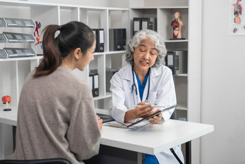Obraz premium An elderly Asian doctor is talking to a younger Asian woman across a desk in a medical office, monthly health check appointment.