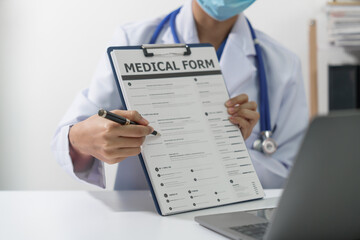 A doctor in a white coat and stethoscope is holding a medical form and a pen, with a laptop in the background. - obrazy, fototapety, plakaty