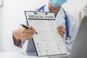 A doctor in a white coat and stethoscope is holding a medical form and a pen, with a laptop in the...