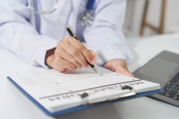 A doctor in a white coat and stethoscope is holding a medical form and a pen, with a laptop in the background.