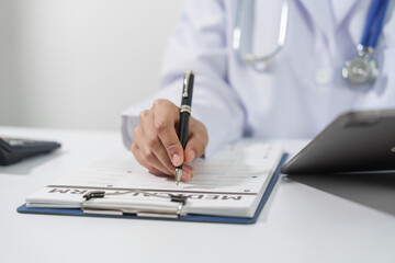 A doctor's hand is writing on a medical record form on a clipboard, with a stethoscope and tablet in the background.