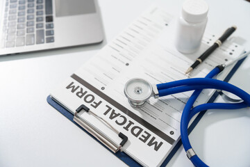 A stethoscope and a pen lie on a medical form on a clipboard, next to a laptop, on a white surface.