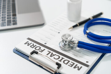 A stethoscope and a pen lie on a medical form on a clipboard, next to a laptop, on a white surface.