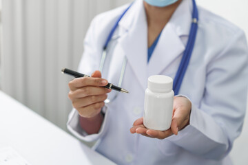 A doctor wearing a face mask is holding a pen and a white medicine bottle, possibly explaining medication instructions.