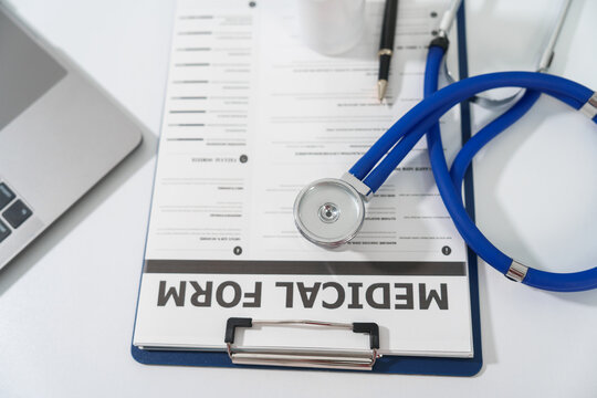 A stethoscope and a pen lie on a medical form on a clipboard, next to a laptop, on a white surface.