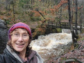 Mature woman takes a selfie near Sgwd Gwladys - one of the many waterfalls near Pontneddfechan in South Wales. She is wearing warm clothing and is wet because of the rain and water spray.
