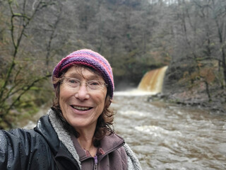 Senior woman taking a selfie at Sgwd Gwladys - one of the many waterfalls near Pontneddfechan in South Wales. She is wearing warm clothing and is wet because of the rain and water spray.