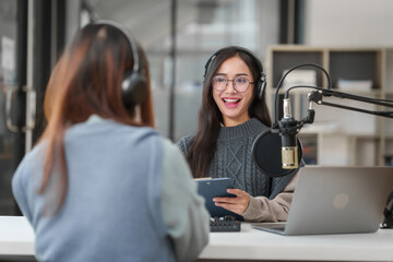Pretty Asian women podcasting. One speaks into microphone and wears headphones, while the other...
