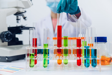 A scientist examines samples with a microscope in a laboratory with test tubes filled with colorful liquids in the background.