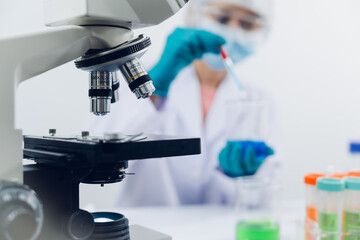 A scientist examines samples with a microscope in a laboratory with test tubes filled with colorful liquids in the background.