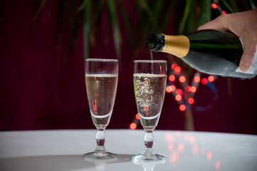 Pouring of brut champagne, cava or prosecco wine in glasses with garland lights on dark background, new year or birthday party