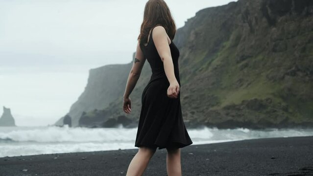 Young beautiful woman in black dress dancing spinning on black sand beach Iceland, slow motion, dramatic waves seascape	