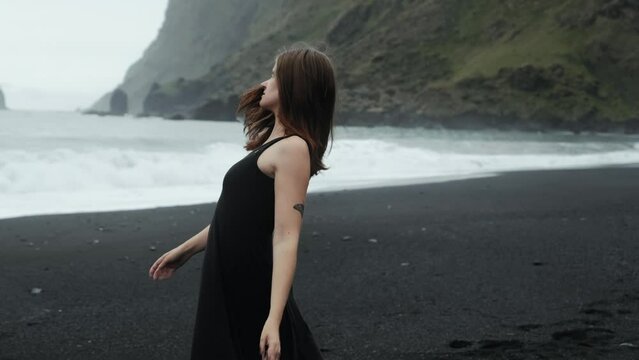 Young beautiful woman in black dress dancing on black sand beach Iceland, slow motion spinning, dramatic waves seascape	