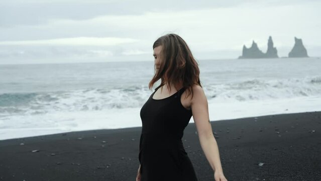 Young beautiful woman in black dress dancing on black sand beach Iceland, dramatic waves seascape, slow motion spinning