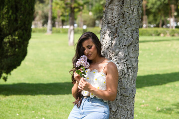 Beautiful Spanish brunette woman with long curly hair and big slit eyes smells a bouquet of violet flowers. The woman is smiling and happy and is leaning against the trunk of a tree in the park.