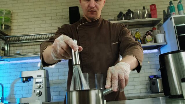 Kitchen worker stirs mixture in saucepan surrounded by professional kitchen equipment. Usage of whisk allows mixing ingredients perfectly