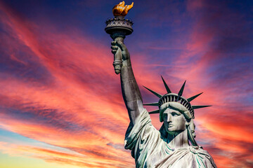 The Statue of Liberty holding her torch on a sunny day, under a clear reddish-orange sky at sunset,...