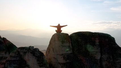Man tourist hiker meditates zen on top of cliff in mountains. Raises his arms to sides, simulating free flight. Peace from nature, enjoying view of mountains at sunset in rays of sunshine. Aerial view