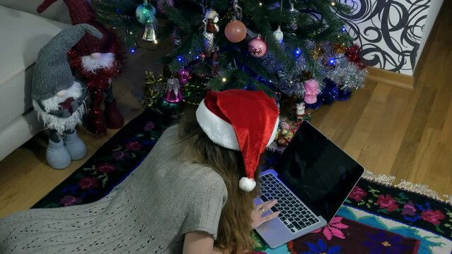 The girl is lying on a mat near a green Christmas tree with a laptop. View of the Christmas tree from the bottom to the top. A girl near a beautiful Christmas tree on a mat with a laptop. High 4k