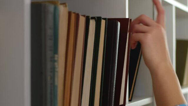 Person carefully taking a book from the row standing on a shelf of a white bookcase at home close up. Concept of curiosity in education and thirst for knowledge