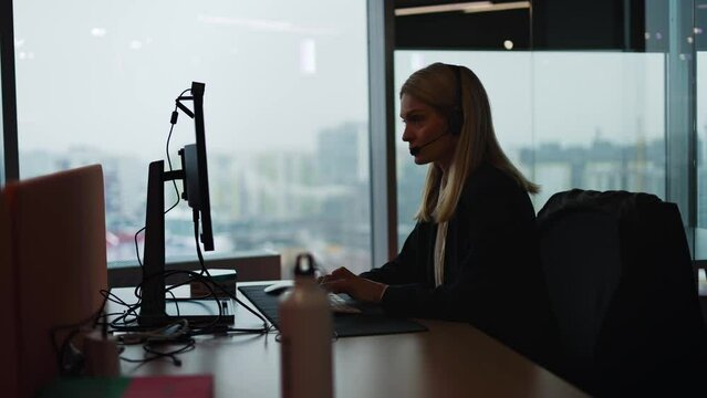 Silhouette view of adult customer support operator assistant working at helpdesk with a headset online from office with panoramic window technical support internet communication headphones microphone.