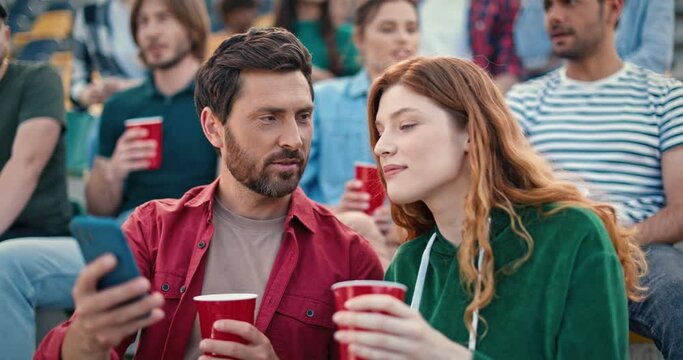 Good-looking Caucasian couple spending time together at football championship. Attractive man showing his lovely woman something on smartphone. People speaking about soccer while holding drink.