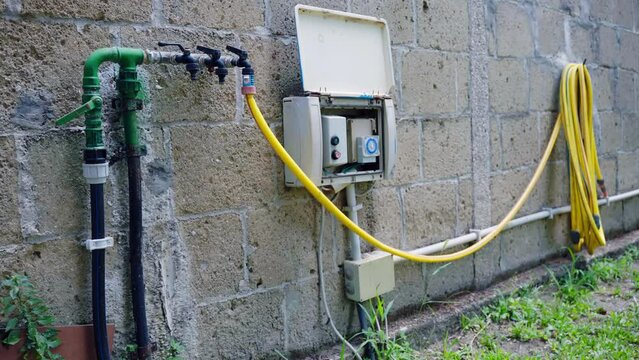 Electrical outlet box with water irrigation indicator installed on the wall, faucet with yellow hose pipes for watering field or garden, pumping water system