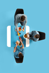 Creative collage. Three employers sitting at table and eating during business lunch against blue studio background. Top view. Concept of business lunch, morning meeting, briefing. Copy space. Ad