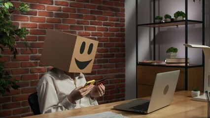 Portrait of female in cardboard box with emoji on head. Worker at the desk holding smartphone and credit card typing number. - obrazy, fototapety, plakaty