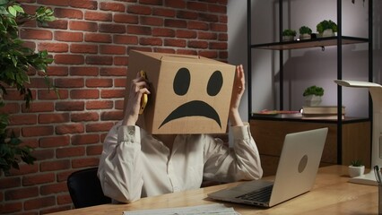 Portrait of a woman in a cardboard box with a negative emoji on her head. Employee sitting at desk,...