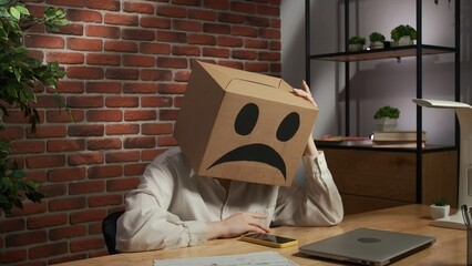 Portrait of female in cardboard box with emoji on head. Worker sitting at the desk looking at smartphone sad hopeless expression.