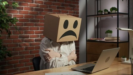 Portrait of female in cardboard box with emoji on head. Worker sitting at the desk working on...