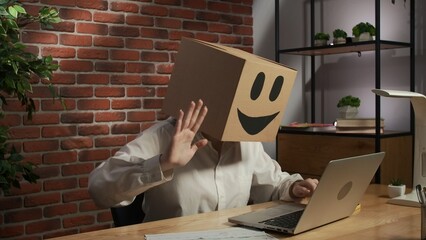 Portrait of female in cardboard box with emoji on head. Worker sitting at the desk talking by video call on laptop, happy face.