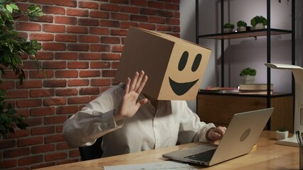 Portrait of female in cardboard box with emoji on head. Worker sitting at the desk talking by video...