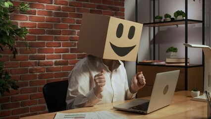 Portrait of a woman in a cardboard box with a smiling smiley face on her head. Employee at desk working on laptop. - obrazy, fototapety, plakaty