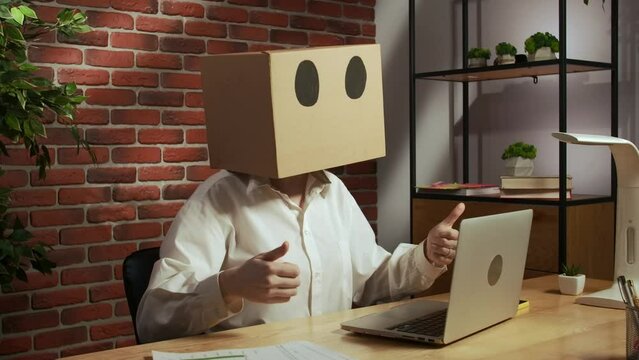 Portrait of female in cardboard box over head. Office worker at the desk working on laptop, winning gestures showing thumbs up.