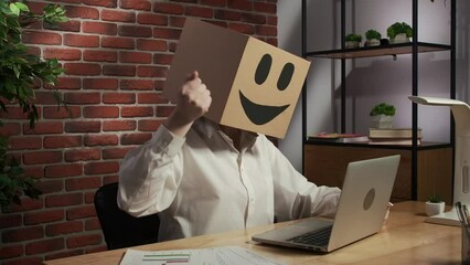 Portrait of female in cardboard box with emoji on head. Worker sitting at the desk working on laptop, happy expression hand up.