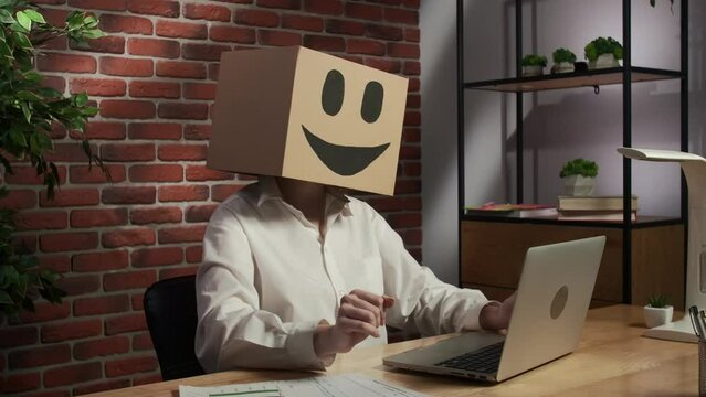 Portrait of female in cardboard box with emoji on head. Worker sitting at the desk working on laptop, leaning back on the chair.
