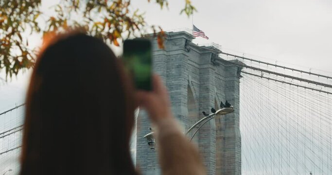 Female tourist taking pictures of the Brooklyn Bridge with an American Flag waving in the wind