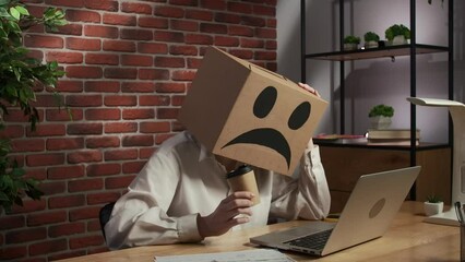 Portrait of female in cardboard box with emoji on head. Worker sitting at the desk holds cup of coffee looks at laptop, sad face.