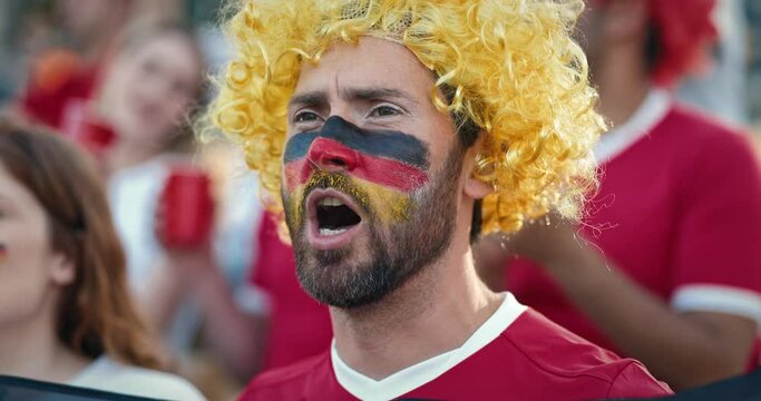 Very close camera view of handsome bearded Caucasian male with yellow wig singing or shouting something in support of his national team. Painted his face with three colours of German national flag.