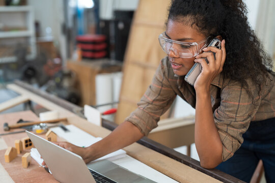 Female carpenter using smart phone and working with laptop computer in wood workshop. Black female carpenter contact to customer by smart phone. SME, Start up and small business concept