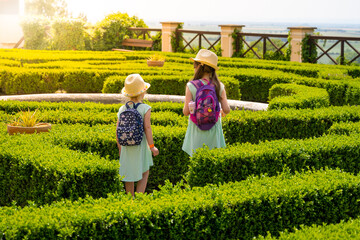 Two small long hair girls in light blue dress with bags and hats walking in the park on sunny...