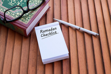 Ramadan Checklist is written on the notepad on the wooden table with a pen, and a holy Quran at the side. Ramadan concept